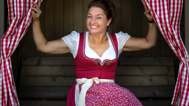 Ursula Weber im rot gekleideten Dirndl blickt zwischen zwei rot, karierten Vorhängen hervor. 