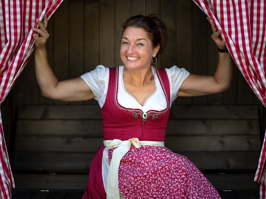 Ursula Weber im rot gekleideten Dirndl blickt zwischen zwei rot, karierten Vorhängen hervor. 