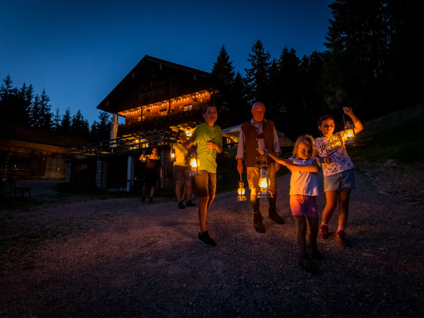 Kinder mit Laternen in der Hand wandern in der Dunkelheit gemeinsam mit dem Wanderführer von der Denkalm ins Tal. 