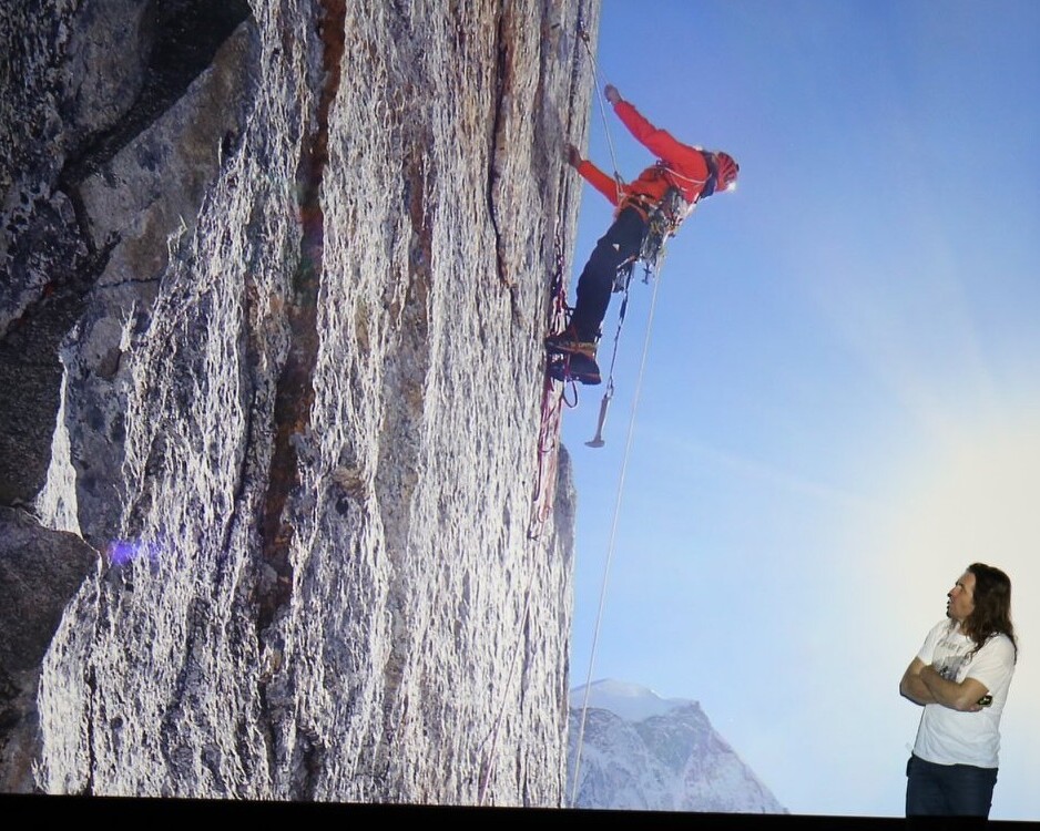 Kletterer Thomas Huber steht vor einer Leinwand, die ihn selber beim Klettern an einer Felsenwand zeigt, und hält seinen Vortrag. 