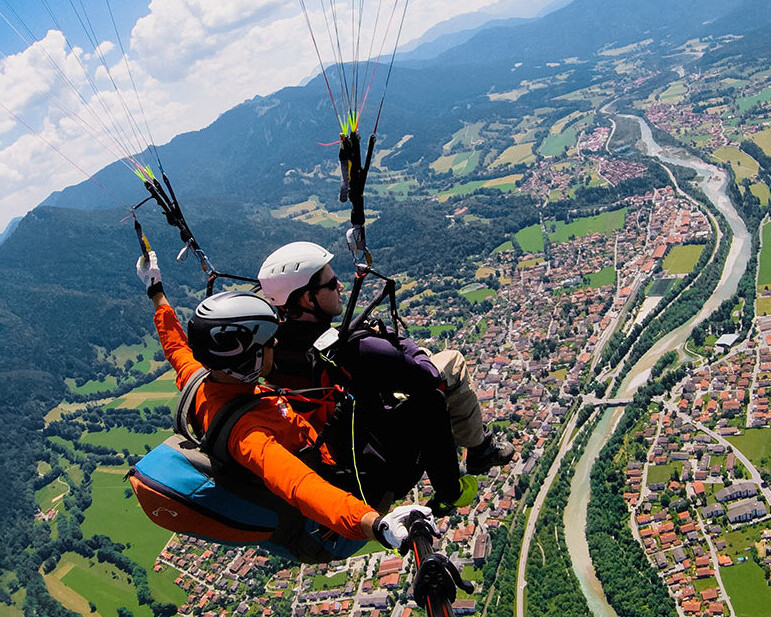 Ein Tandemflieger fliegt mit seinem Gleitschirm, der mit zwei Personen besetzt ist, bei Sonnenschein und blauen Himmel über Lenggries. Man sieht das Dorf Lenggries mit den Häuser, Wiesen, Felder und der Isar von der Vogelperspektive. 