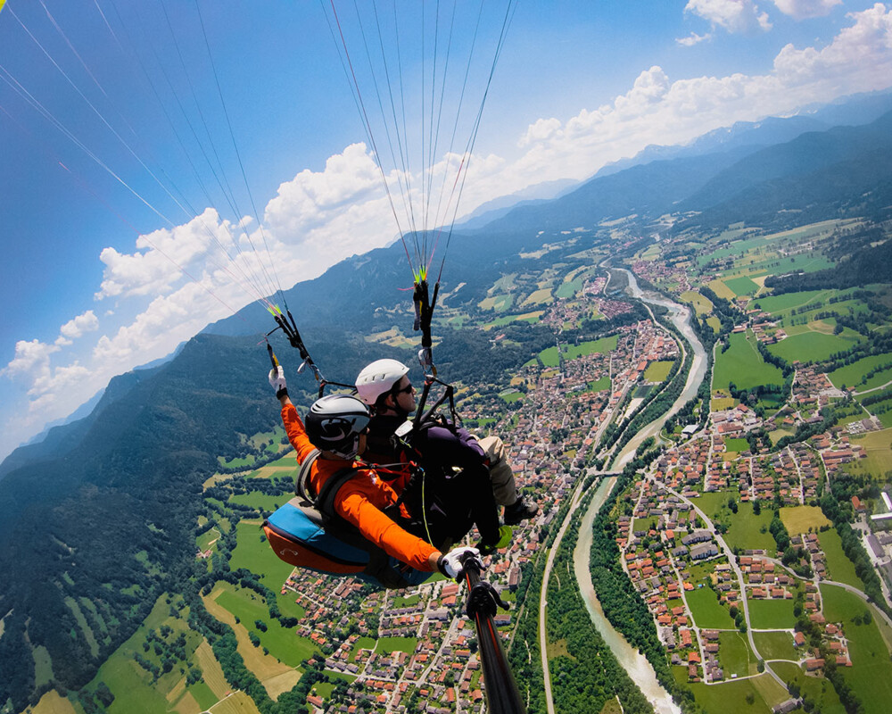 Ein Tandemflieger fliegt mit seinem Gleitschirm, der mit zwei Personen besetzt ist, bei Sonnenschein und blauen Himmel über Lenggries. Man sieht das Dorf Lenggries mit den Häuser, Wiesen, Felder und der Isar von der Vogelperspektive. 